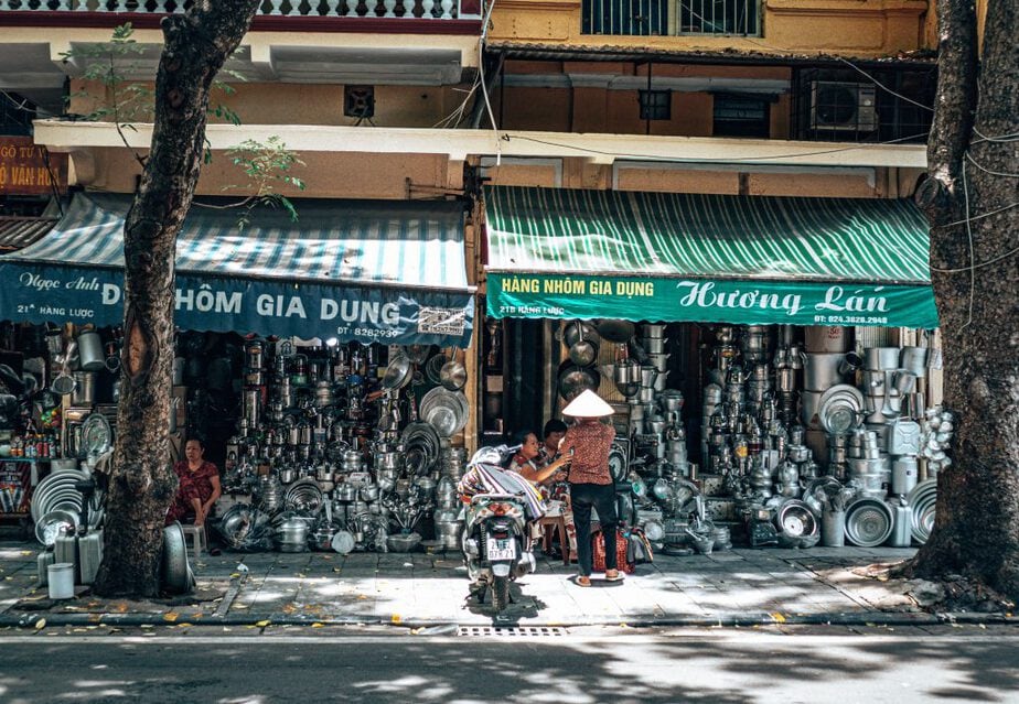 Kitchenware stores with load of aluminum kitchenware and a person wearing Vietnamese traditional hat standing in front of the stores and beside his scooter