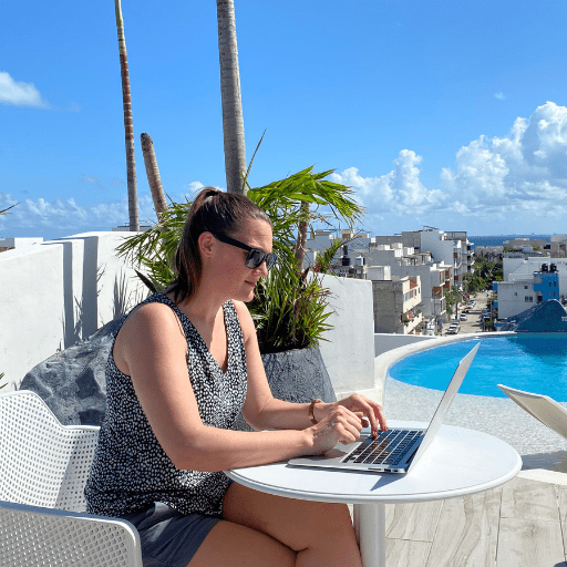 A woman working on a laptop at a small white table with a pool and blue skies in the background